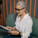Elderly woman in glasses reviewing documents on clipboard, sitting indoors with a focused expression.